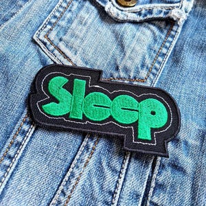 Può includere: Una toppa nera con la scritta "Sleep" in verde brillante, bordata di bianco. La toppa è cucita su una giacca di jeans blu chiaro, con cuciture visibili e un dettaglio di tasca.