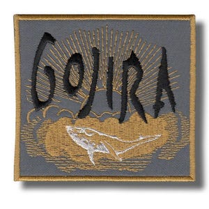 Może przedstawiać: Haftowana naszywka z napisem "GOJIRA" w czarnym skrypcie nad złotym i białym wzorem wieloryba. Naszywka ma szare tło ze złotą obwódką i wzorem promieni słonecznych.