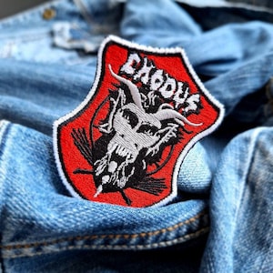 May include: A red, white, and black Exodus patch with a shield shape. The patch features a detailed illustration of a skull with horns and the band's name in white block letters. The patch is set against a blue denim jacket.