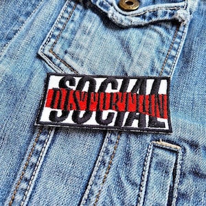 Könnte beinhalten: Ein rechteckiger Aufnäher mit den Worten "SOCIAL DISTORTION" in Schwarz und Rot auf weißem Grund gestickt. Der Aufnäher ist auf eine hellblaue Jeansjacke, in der Nähe der Tasche, genäht.