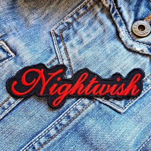 Könnte beinhalten: Ein schwarzer, gestickter Aufnäher mit dem Wort "Nightwish" in roter Schrift. Der Aufnäher ist auf eine hellblaue Jeansjacke genäht. Die Jacke hat sichtbare Nähte und einen Metallknopf.