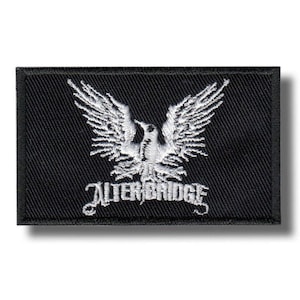Op de afbeelding: Zwart rechthoekig embleem met een wit geborduurde adelaar met uitgespreide vleugels en de tekst "ALTER BRIDGE". De patch heeft een zwarte rand en een witte schaduw.