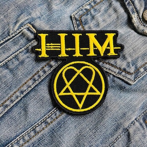 Puede incluir: Parche bordado en negro y amarillo con las letras "HIM" sobre un emblema circular con un pentagrama en forma de corazón. El parche está cosido a una chaqueta vaquera azul.