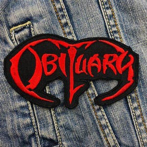 May include: Black and red embroidered patch with the word "OBITUARY" in a stylized font. The patch is rectangular with rounded edges and is set against a denim background.