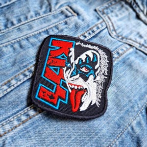 May include: Embroidered patch with the word "KISS" in red letters with a blue outline, and a detailed illustration of a band member's face. The patch is black with white, blue, and red accents, and is sewn onto a denim jacket.