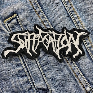 Può includere: Toppa ricamata in bianco e nero con la scritta "STARVATION" in un carattere stilizzato. La toppa è cucita su una giacca di jeans blu, con cuciture e trama del tessuto visibili.