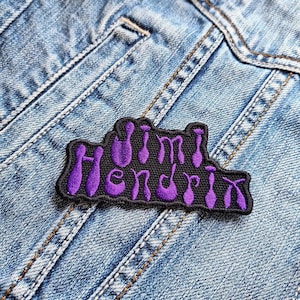 Könnte beinhalten: Ein schwarzer Aufnäher mit dem Namen "Jimi Hendrix" in lila Schrift gestickt. Der Aufnäher ist auf eine hellblaue Jeansjacke genäht, mit sichtbaren Nähten und Taschen.