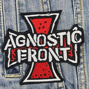 Puede incluir: Parche bordado con el texto "AGNOSTIC FRONT" en letras blancas sobre un diseño en forma de cruz negra con un centro rojo. El parche está cosido en una chaqueta vaquera.