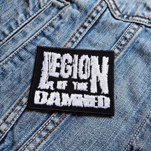 Op de afbeelding: Een zwart rechthoekig embleem met witte geborduurde tekst "LEGION OF THE DAMNED". De patch is op een lichtblauwe spijkerjas genaaid. De patch is een kledingaccessoire.