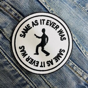 May include: A circular white embroidered patch featuring the text "SAME AS IT EVER WAS" in black. A black silhouette of a person walking is in the centre. The patch is set against a blue denim jacket.