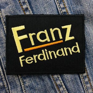 Könnte beinhalten: Schwarzer, gestickter Aufnäher mit dem Text "Franz Ferdinand" in gelbem Garn. Das Wort "Franz" steht über "Ferdinand", getrennt durch einen orangefarbenen Strich. Der Aufnäher ist rechteckig und vor einem Jeans-Hintergrund platziert.