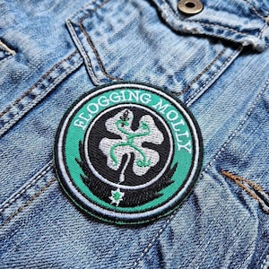 Könnte beinhalten: Ein runder Aufnäher mit dem Text "Flogging Molly" in weißer Schrift auf einem grünen Rand. Der Aufnäher zeigt ein weißes und grünes Kleeblatt-Design auf schwarzem Hintergrund, vor einer Jeansjacke.
