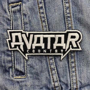 Può includere: Toppa ricamata in bianco e nero con la scritta "AVATAR COUNTRY" su una giacca di jeans blu. La toppa ha un carattere audace e stilizzato e un bordo bianco. La giacca di jeans ha cuciture visibili.