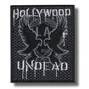 Puede incluir: Parche bordado en negro y gris con las palabras "HOLLYWOOD UNDEAD" y "LA" y un par de alas estilizadas. El parche tiene un fondo texturizado y un estilo de fuente goteante.