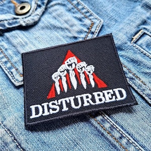 May include: A black square patch featuring the word "DISTURBED" in white, bold lettering. Above the word is a red triangle with white stylized figures. The patch is on a denim jacket.