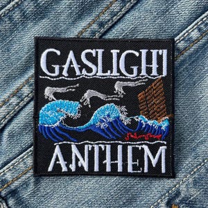 Op de afbeelding: Zwart vierkantige patch met de woorden "GASLIGHT ANTHEM" in wit, gebogen aan de boven- en onderkant. De patch heeft een blauw golfontwerp met grijze handen en een bruine structuur.
