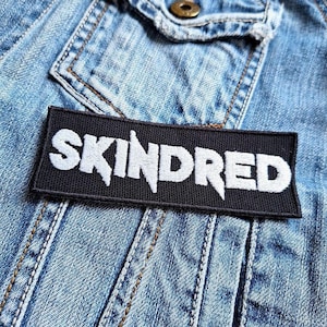 Peut inclure: Un écusson rectangulaire noir avec le mot "SKINDRED" en blanc, avec des lettres dentelées. L'écusson est cousu sur une veste en jean bleu clair, avec des coutures visibles et un bouton.