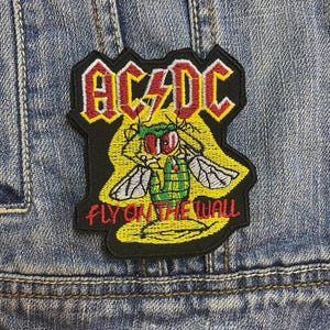 May include: Embroidered patch with the AC/DC logo in red and yellow, and a cartoon fly illustration. The text "FLY ON THE WALL" is below the fly. The patch is attached to a denim jacket.