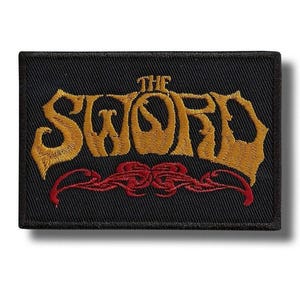 May include: Black rectangular patch with the text "THE SWORD" in gold, stylized lettering. Below the text is a red decorative design. The patch has a black border and is likely an embroidered patch.