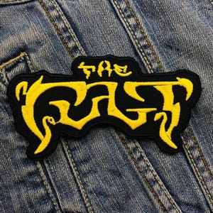 Peut inclure: Un écusson noir avec le texte "THE CULT" brodé en jaune. L'écusson, de forme incurvée, est posé sur un fond en denim bleu.