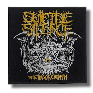 Pode incluir: Patch quadrado preto com o nome da banda "SUICIDE SILENCE" em letras amarelas estilizadas. Abaixo, um gráfico detalhado em branco e cinza com um olho em um triângulo. O texto "THE BLACK CROWN" está na parte inferior em amarelo.