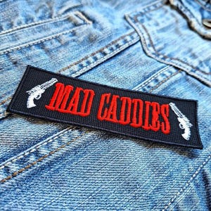 Op de afbeelding: Een zwart rechthoekig embleem met de woorden "MAD CADDIES" in het rood geborduurd, geflankeerd door witte pistoolontwerpen. Het embleem is genaaid op een lichtblauwe spijkerjas, waarbij de textuur van de stof zichtbaar is.