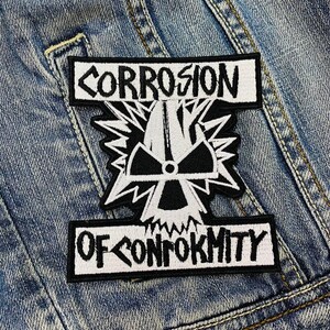 May include: A black and white embroidered patch with the words "CORROSION" and "OF CONFORMITY" in white block letters. The patch features a central graphic of a stylized radiation symbol within a starburst design, set against a denim jacket.