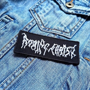 Könnte beinhalten: Ein schwarzer rechteckiger Aufnäher mit weiß gesticktem Text, platziert auf einer blauen Jeansjacke. Der Text scheint ein Bandname oder -logo zu sein. Der Aufnäher ist ein potenzielles Accessoire für Kleidung.