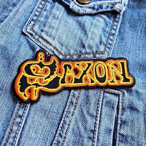 Può includere: Toppa ricamata nera e arancione con la scritta "NIXON" in un carattere stilizzato. La toppa è cucita su una giacca di jeans blu chiaro, con cuciture e dettagli delle tasche visibili.