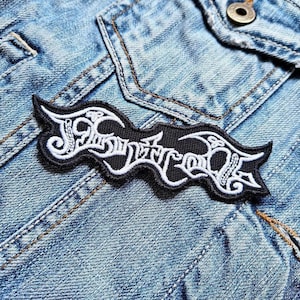 Puede incluir: Un parche bordado en blanco y negro con la palabra "Justice" en una fuente estilizada. El parche es rectangular con bordes curvos. El parche está colocado sobre una chaqueta vaquera azul.