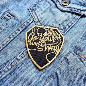 Könnte beinhalten: Ein schwarz-goldener, gestickter Aufnäher mit dem Text "Go Your Own Way" in Schreibschrift. Der Aufnäher zeigt ein Bergdesign und ist auf eine hellblaue Jeansjacke genäht.
