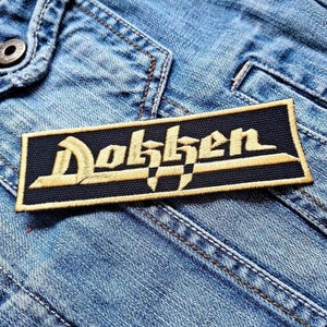 Op de afbeelding: Een rechthoekige patch met het woord "Dokken" geborduurd in goudkleurig garen op een zwarte achtergrond. De patch is genaaid op een lichtblauwe spijkerjas, met zichtbare stiksels en een knoop.