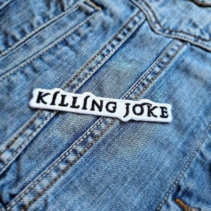 May include: A white patch with the text "KILLING JOKE" in black lettering is sewn onto a blue denim jacket. The patch is rectangular with a slightly irregular border. The denim jacket has visible stitching and pockets.