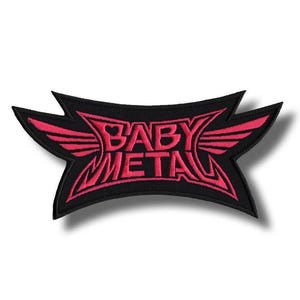 May include: A black patch featuring the pink text "BABY METAL" with a wing design. The patch has a distinctive shape with pointed edges and a curved banner design, suitable for clothing or accessories.