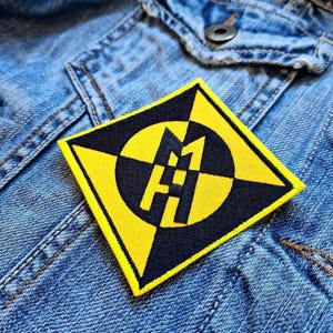 Può includere: Una toppa ricamata gialla e nera di forma quadrata, con un design geometrico e le lettere "M" e "H" al centro. La toppa, con un motivo a scacchiera, è cucita su una giacca di jeans.
