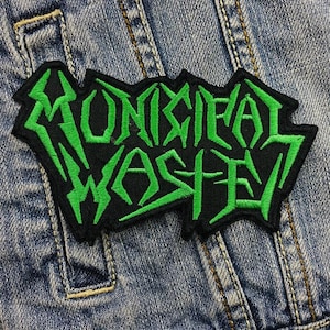 Könnte beinhalten: Ein schwarzer Aufnäher mit dem Schriftzug "MONSTRIAL WASTE" in leuchtend grünen, kantigen Buchstaben. Der Aufnäher ist vor einem blauen Jeans-Hintergrund platziert, der den Kontrast zwischen den Farben und dem Design hervorhebt.