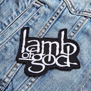 Op de afbeelding: Zwart geborduurde patch met de witte tekst "Lamb of God", genaaid op een lichtblauwe spijkerjas. De patch heeft een zwarte rand en een gestileerd lettertype.