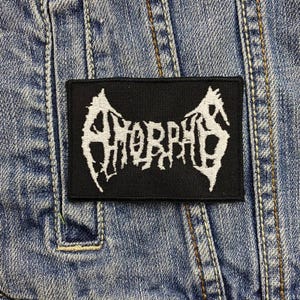 May include: A black rectangular patch with the word "AHORRAS" in white, stylized, gothic lettering. The patch is sewn onto a blue denim jacket. The lettering has a jagged, distressed appearance.