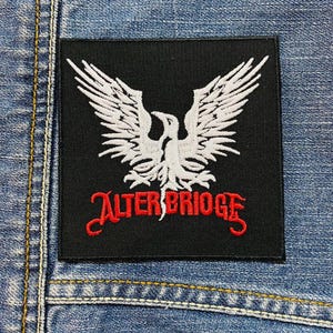 Op de afbeelding: Zwart vierkantige patch met een wit adelaarsontwerp en de rode tekst "ALTER BRIDGE". De patch is op een blauw spijkerjasje genaaid. De adelaar heeft uitgespreide vleugels.