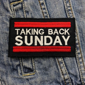 Pode incluir: Um patch retangular preto com as palavras "TAKING BACK SUNDAY" bordadas em branco. O texto é emoldurado por duas listras horizontais vermelhas. O patch é costurado em uma jaqueta jeans.