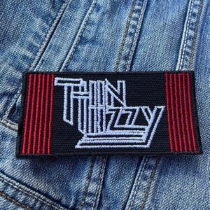 May include: Black rectangular patch with the words "Thin Lizzy" in white, stylized lettering. The patch has red vertical stripes on each end. The patch is on a blue denim background.