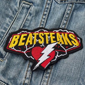 May include: Embroidered patch with the text "BEATSTEAKS" in yellow letters outlined in black and red. The patch features a red heart with a white lightning bolt. The patch is sewn onto a denim jacket.