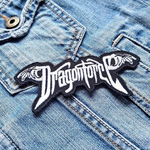 Może przedstawiać: Czarno-biała, haftowana naszywka z napisem "Dragonforce" w stylizowanej czcionce. Naszywka ma motywy przypominające skrzydła na każdym końcu. Naszywka jest przyszyta do jasnoniebieskiej kurtki dżinsowej.