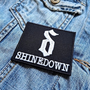 Può includere: Una toppa quadrata nera con la scritta "SHINEDOWN" ricamata in bianco, insieme a un simbolo "S" bianco stilizzato. La toppa è posta su una giacca di jeans blu.