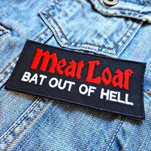 Op de afbeelding: Een rechthoekige zwarte patch met de woorden "Meat Loaf" in rood en "BAT OUT OF HELL" in wit. De patch is genaaid op een lichtblauwe spijkerjas.