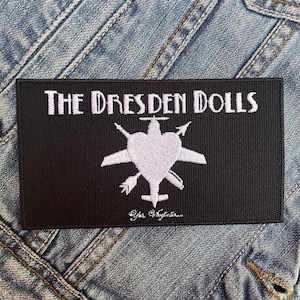May include: Black rectangular patch with white embroidered text "THE DRESDEN DOLLS" above a white heart-shaped design with wings, arrows, and a plane. The text "Yes Virginia..." is below the design. The patch is on a denim jacket.