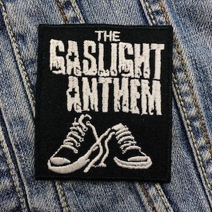 Pode incluir: Patch quadrado preto com o texto "THE GASLIGHT ANTHEM" em branco, com letras desgastadas. Abaixo, dois tênis brancos e pretos com cadarços cruzados. O patch está em um fundo de ganga azul.