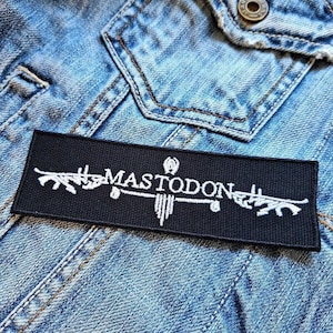 May include: A black rectangular patch with the word "MASTODON" in white, stylized text. The patch is embroidered with white decorative elements on a denim jacket.