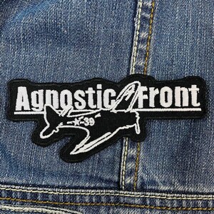 Puede incluir: Parche bordado en blanco y negro con el texto "Agnostic Front" y un diseño estilizado de avión. El parche es rectangular con un borde negro y está cosido a una chaqueta vaquera.