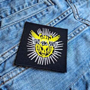 May include: A square, black patch with white and yellow embroidered design. The design features a yellow skull in the center, surrounded by white rays and the words "KILL SWITCH ENGAGE" in white. The patch is placed on a blue denim jacket.